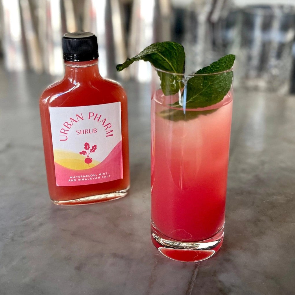 a highball glass filled with reddish pink beverage and garnished with fresh mint alongside a bottle of Urban Pharm Watermelon Mint Shrub