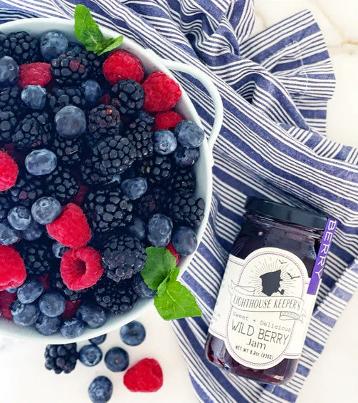 a jar of wild berry jam next to a bowl overflowing with fresh blackberries, raspberries, blueberries laid out on a blue and white striped cloth
