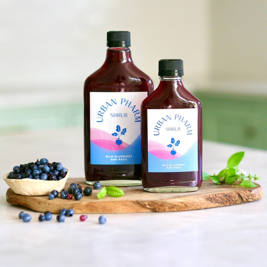 a bottle each of the 200ml and 375ml wild blueberry shrub on a wood board on a countertop alongside fresh blueberries and basil leaves