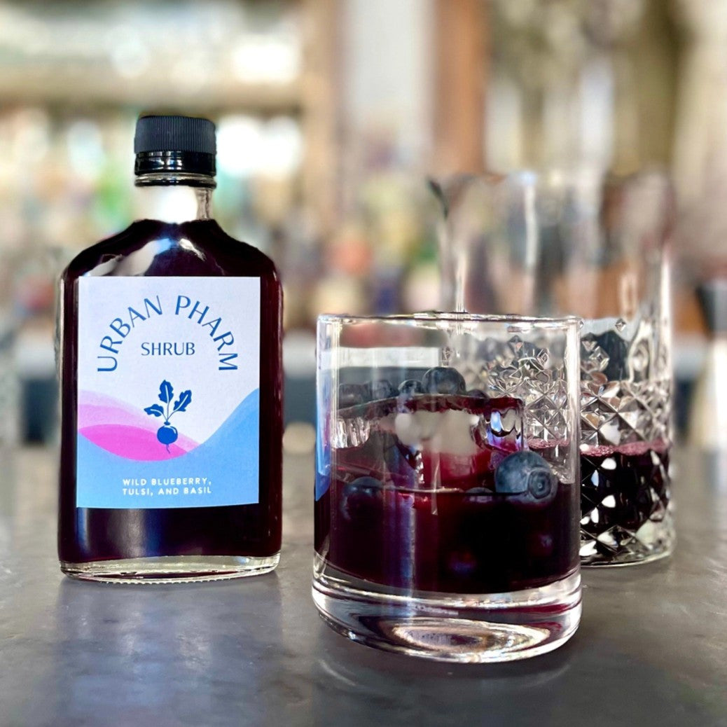 a bar counter with a bottle of Wild Blueberry shrub and two rocks glasses filled with deep purple beverages garnished with blueberries