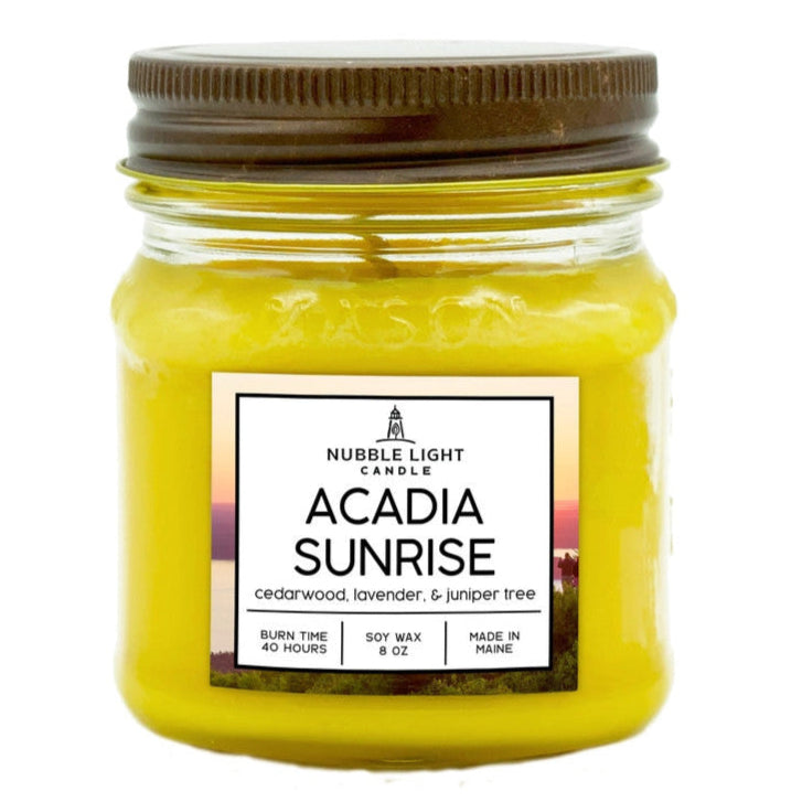 a candle in a clear glass mason jar with bright yellow wax and a label that reads "Acadia Sunrise" against a white background