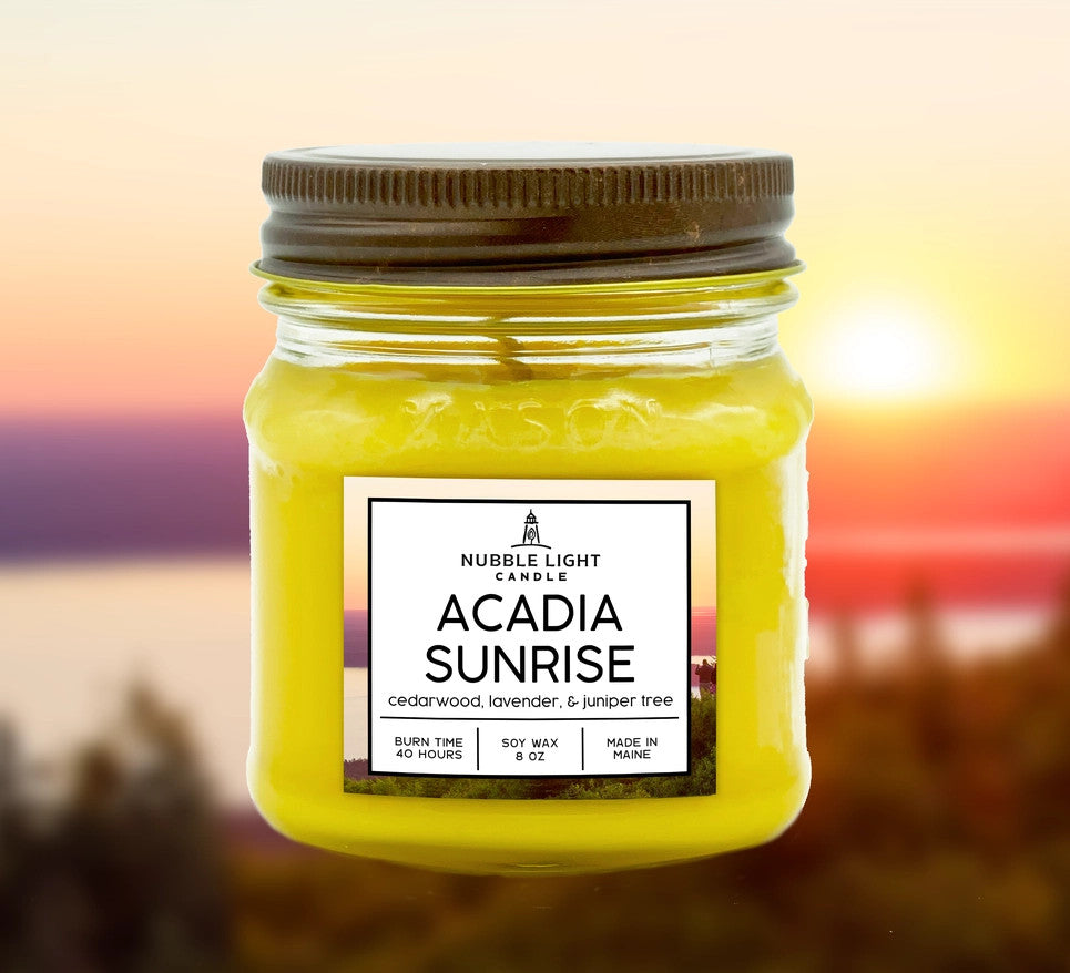 a candle in a clear glass mason jar with bright yellow wax and a label that reads "Acadia Sunrise" with an image of an ocean sunrise behind it
