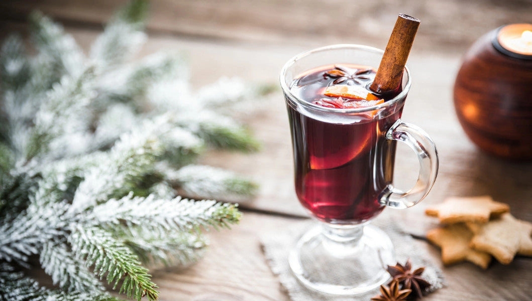 a wintry themed tabletop with balsam boughs and an elegant clear glass mug containing a cranberry-red  mulled beverage, garnished with a citrus, spices, and cinnamon stick