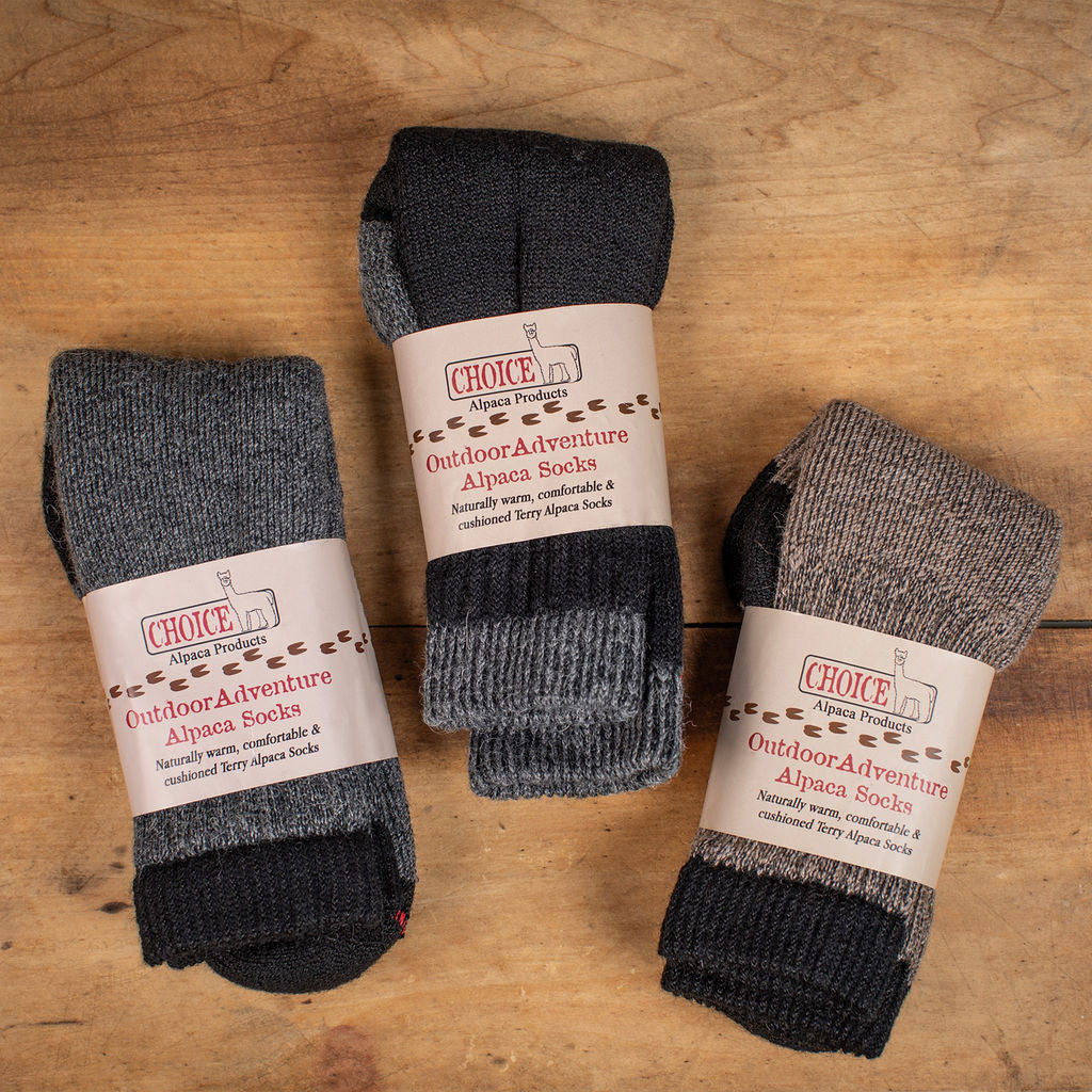 three pairs of alpaca outdoor adventure socks folded in their packaging on a wood background showing the available colors for the socks - gray, black, and cocoa brown