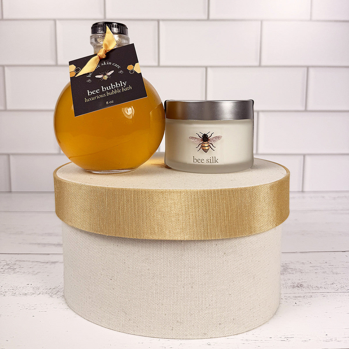 the Bee Bubbly bottle and Bee Silk jar atop the hat box on a white wood surface with white tiled background