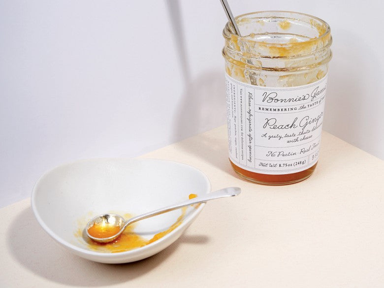 A nearly empty jar of Bonnie's Peach Ginger jam next to a white bowl with a silver spoon coated in jam