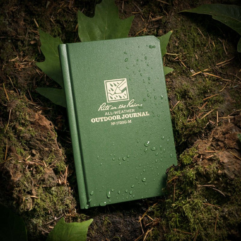 a green hard cover notebook with rain beading on the cover rests on a mossy, muddy forest floor
