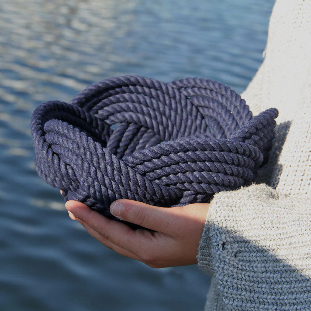 a hand woven cotton rope bowl in navy blue seen in profile in the hands of someone standing at the waterfront