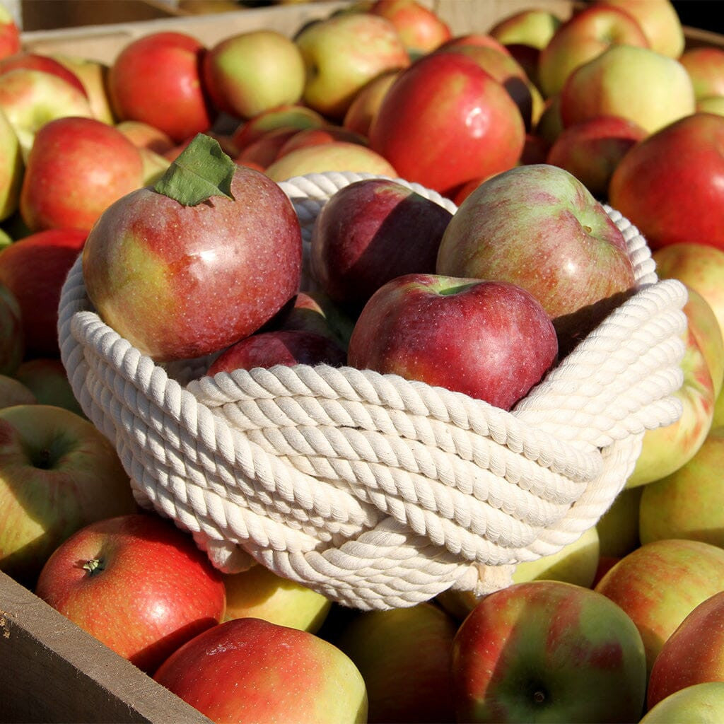 a hand woven white cotton rope bowl filled with 5 or 6 large apples resting on a bed of apples