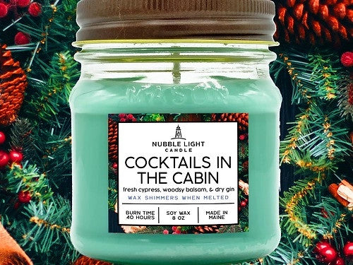 a candle in a clear glass mason jar with light green wax and a label that reads "Cocktails in the Cabin" against a festive background of pine boughs, pinecones, and winterberries