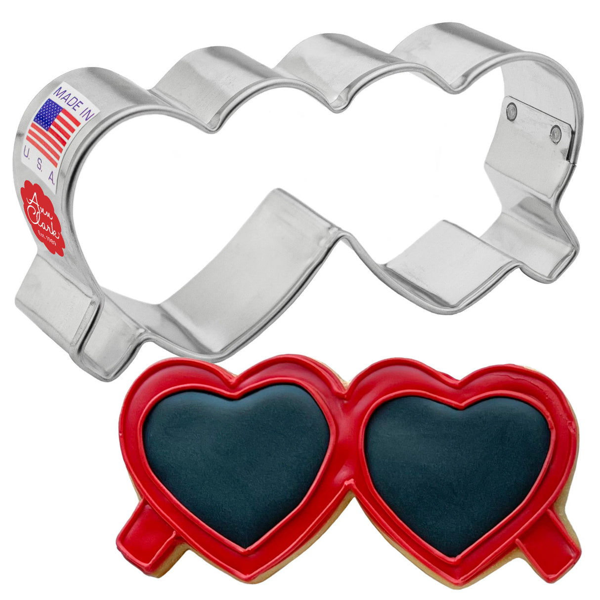 a silver, tin cookie cutter in the shape of folded heart-shaped glasses next to a fully decorated cookie of folded heart shaped sunglasses. the frames are decorated with red icing and the lenses of the glasses have black icing.
