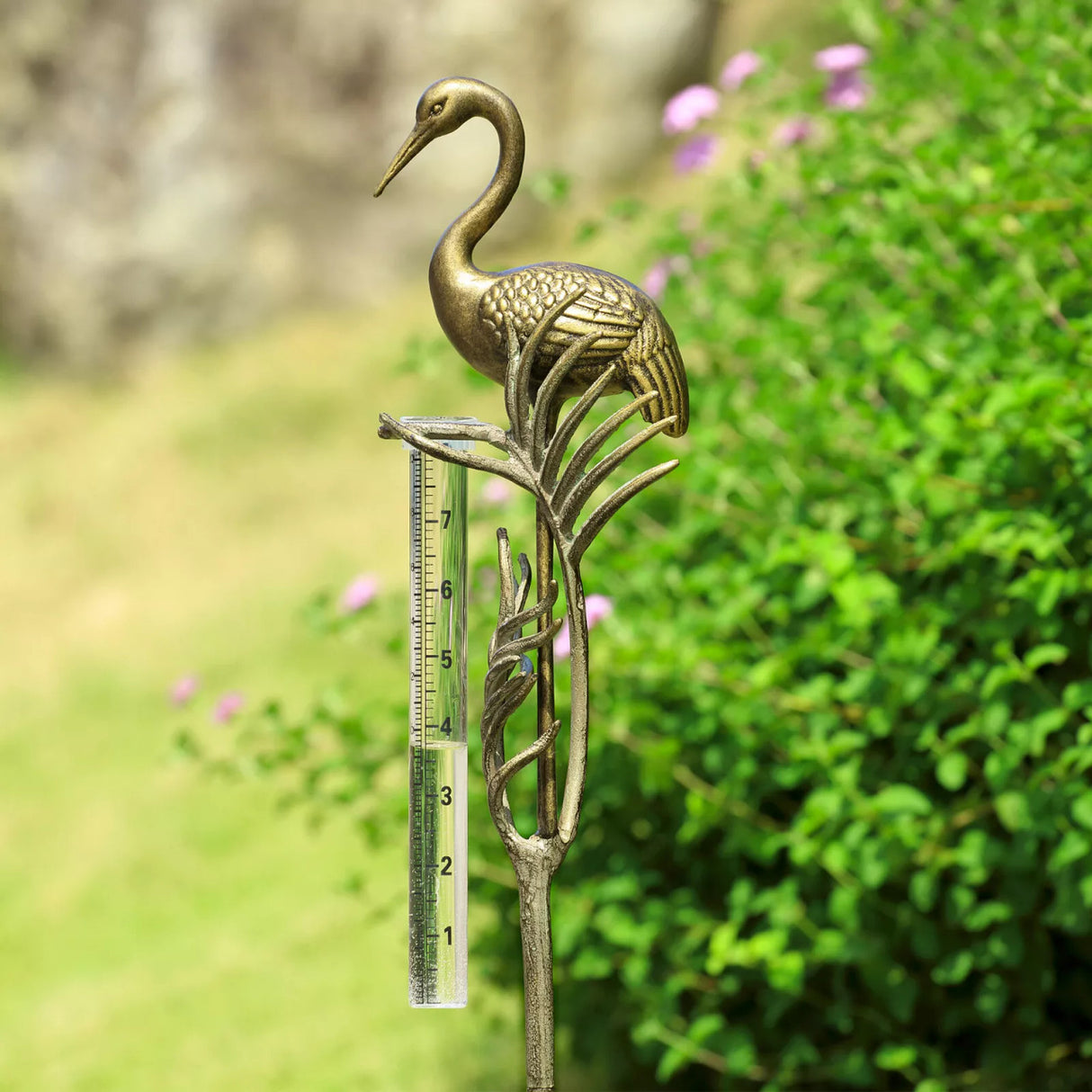 Medium close up view of the crane rain gauge in a garden, featuring the elegant details of the crane, sculpted fronds, and clear rain gauge with markings to measure rainfall
