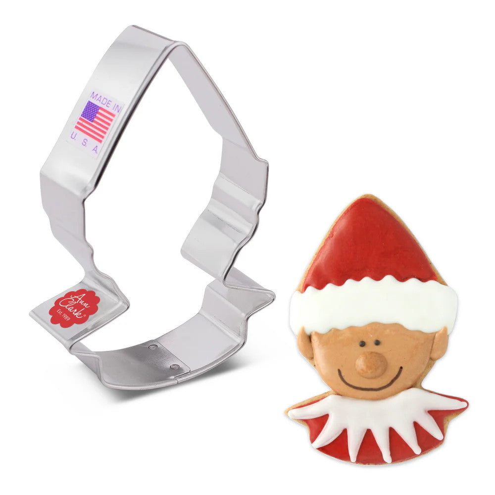 A tin cookie cutter in the shape of an elf's head with a pointed elf hat next to a fully decorated cookie in the shape of an elf's head with a pointed hat iced with red icing and white trim on the hat, a smiling elf face with medium skin tone, and a white pointed collar on a red shirt