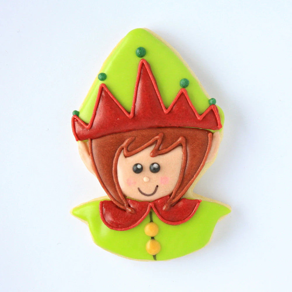 a fully decorated cookie in the shape of an elf's head with a pointed hat iced with green icing and red decorative detail on the hat, a smiling elf face with fair skin tone, and a red, rounded collar on a green shirt