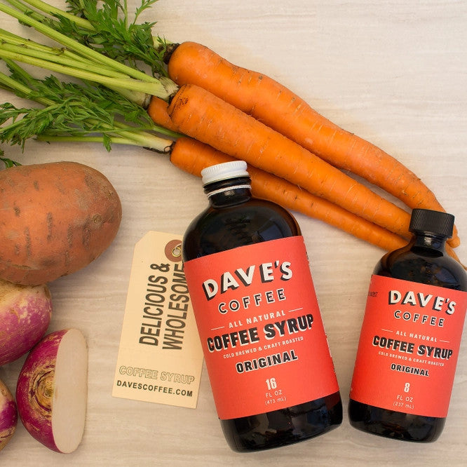 two bottles of Dave's coffee syrup laying on a countertop next to fresh carrots and onions, portraying the syrup as a New England grocery staple