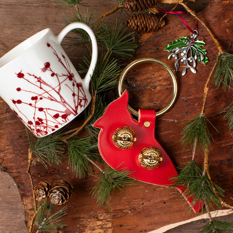 the gifts included in the Festive Accents gift basket laid out on a festive bark background with evergreen and pinecones - the winterberries mug, the cardinal bell door hanger, and the mistletoe ornament
