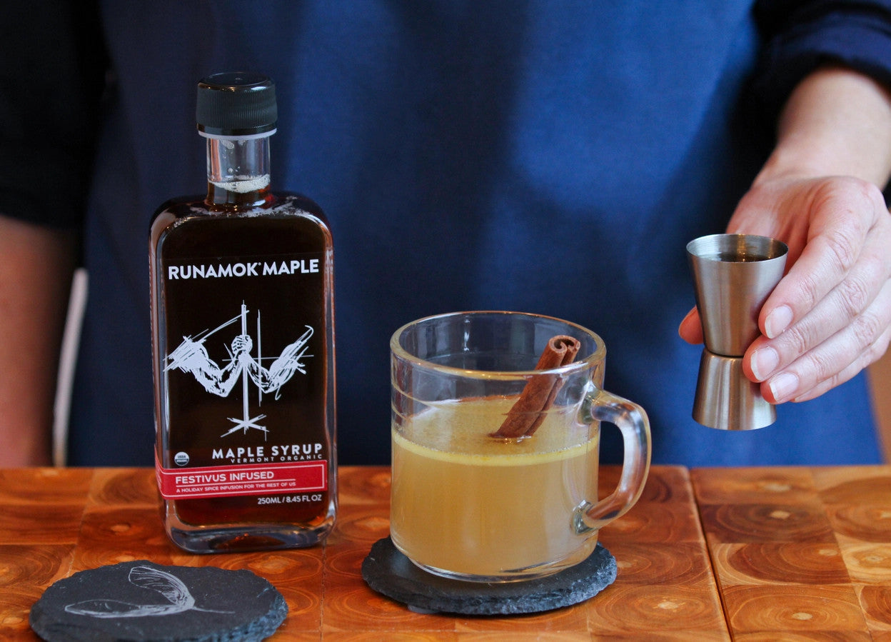 a bar scene with a drink being mixed - a bottle of festivus infused maple syrup sits on the bar next to a rocks glass filled with a cider like beverage and a cinnamon stick, showing that the syrup has versatile use for cocktails