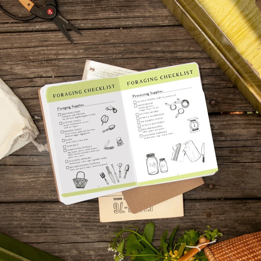 The foraging guide book open to a checklist page on a rustic wood table with scissors and a basket of foraged plants
