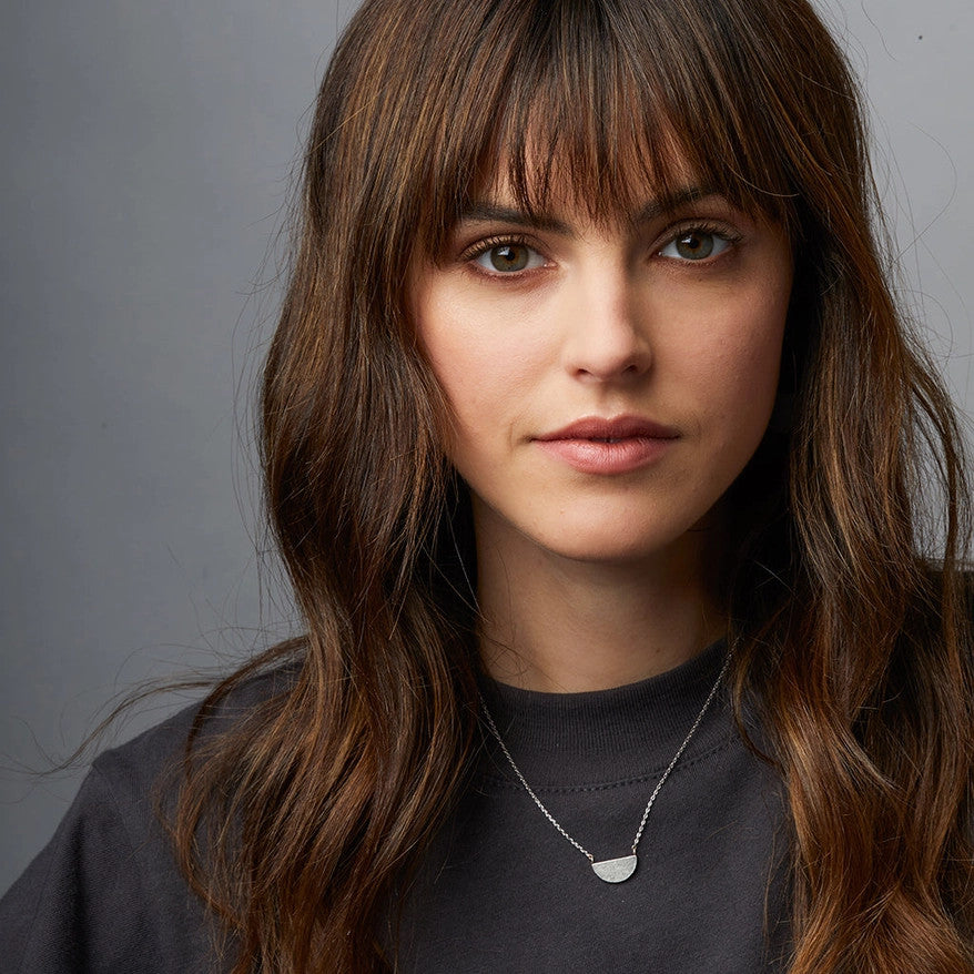 A silver necklace with a half moon pendant seen on a model showing the scale of the necklace and the chain length - the pendant lands just below the collarbone