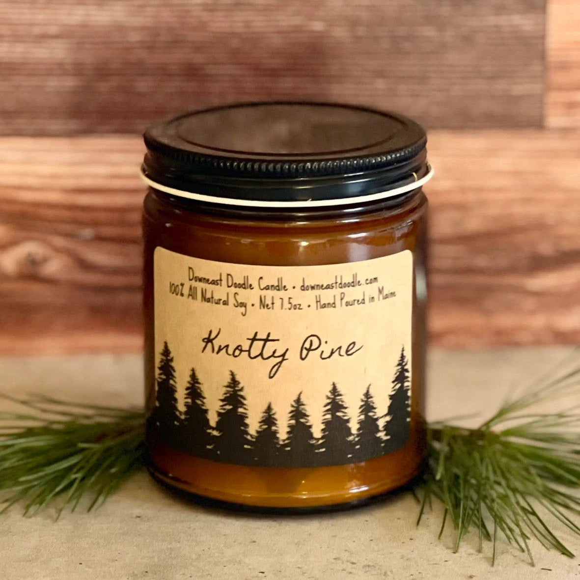 a candle in an amber glass jar with black lid and kraft label with handwriting style graphics that read "Knotty Pine" above a silhouetted forest of pine trees flanked by evergreen sprigs