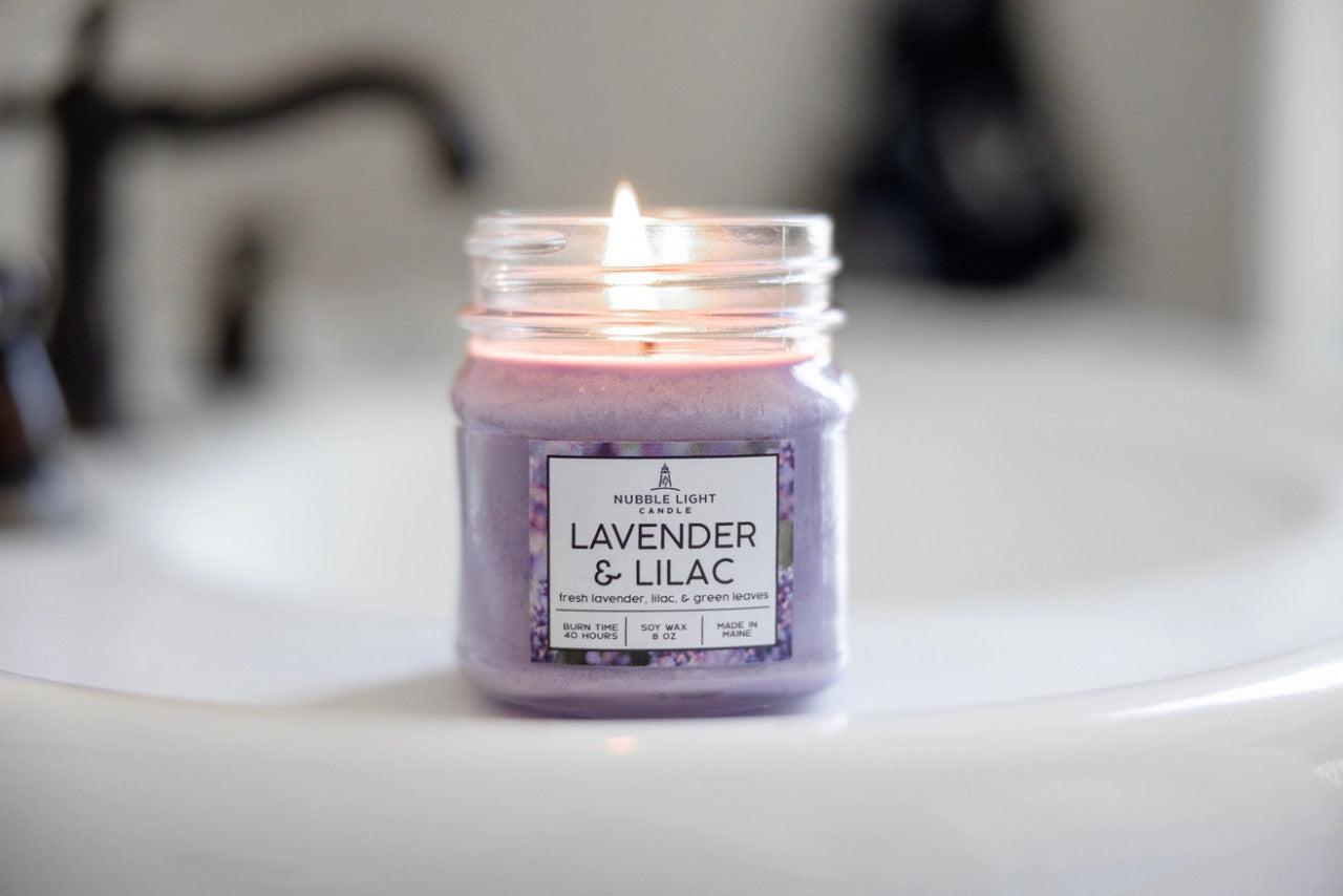a candle in a clear glass mason jar with light purple wax and a label that reads "Lilac & Lavender" on a bathroom sink