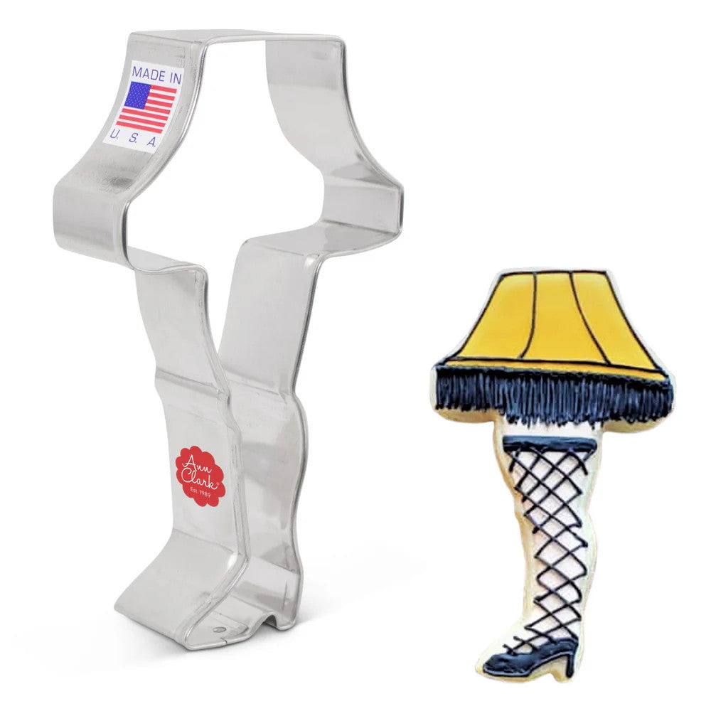 a tin cookie cutter in the shape of a leg in high heels topped with a lampshade next to a fully decorated cookie in the same shape and frosted with yellow icing on the lampshade with black fringe, a black high heeled shoe on the leg, and fishnet stockings on the leg