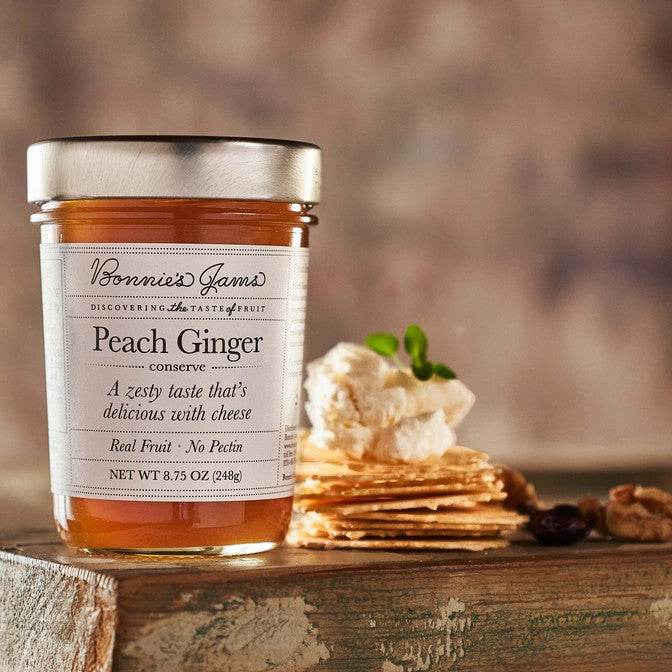 Bonnie's Peach Ginger jam jar with an elegant white label and silver jar top on a table next to a stack of crackers topped with a soft cheese and herbs