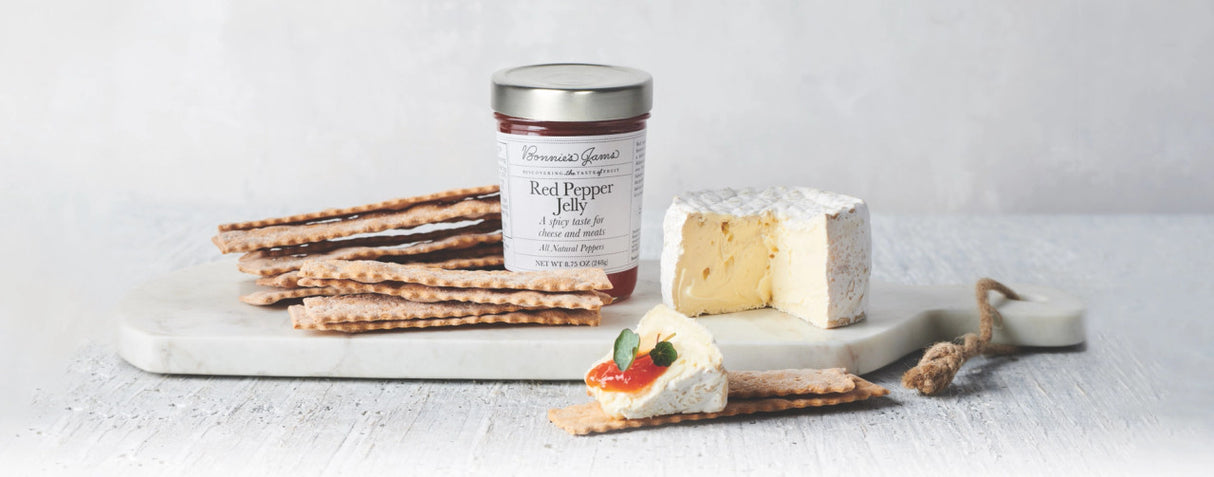 A white rustic tabletop with a white marble cheese board topped with a jar of Bonnie's red pepper jelly, flatbread crackers, a small wheel of soft cheese, and a cracker topped with cheese and red pepper jelly