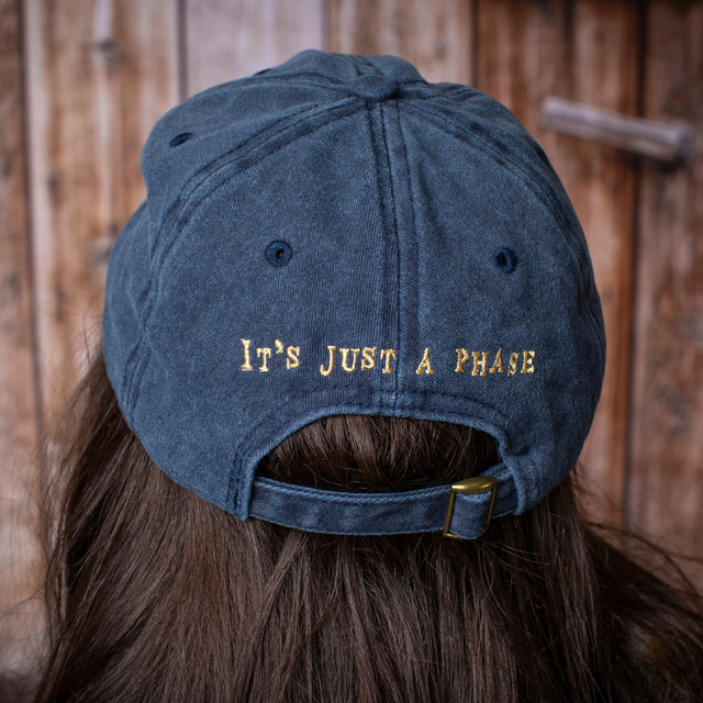 a model wearing the baseball cap showing the embroidered detail on the back of the cap that reads "It's just a phase"