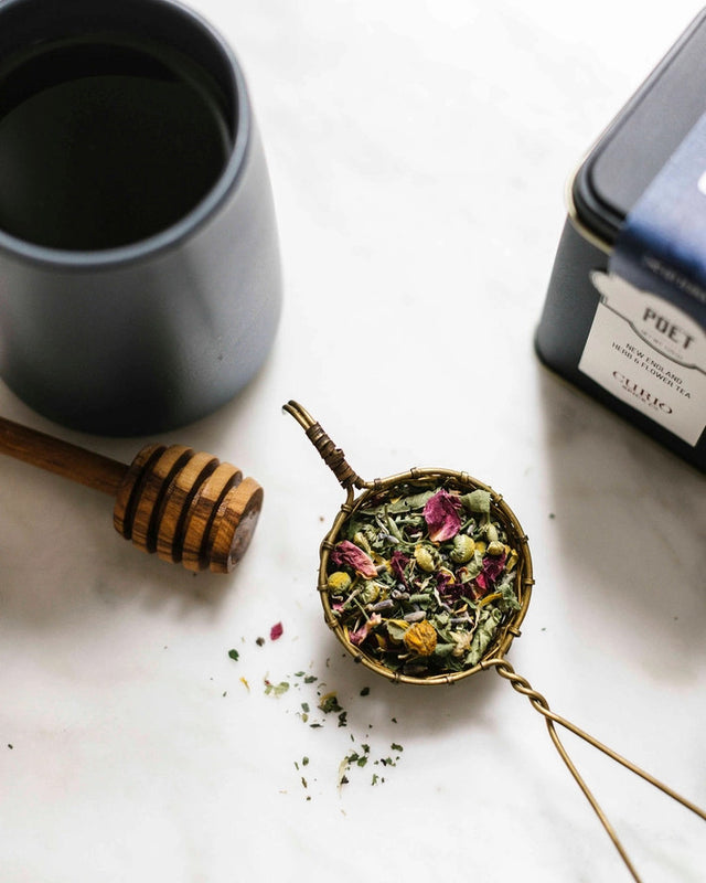 a tea infuser filled with a colorful floral tea next to the tea tin, a storage pot, and a honey server