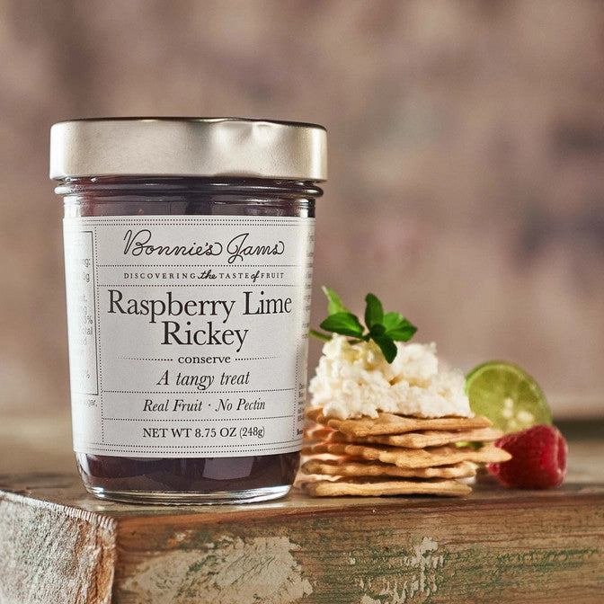 Bonnie's Raspberry Lime Rickey jam jar with an elegant white label and silver jar top on a table next to a stack of crackers topped with a soft cheese and herbs