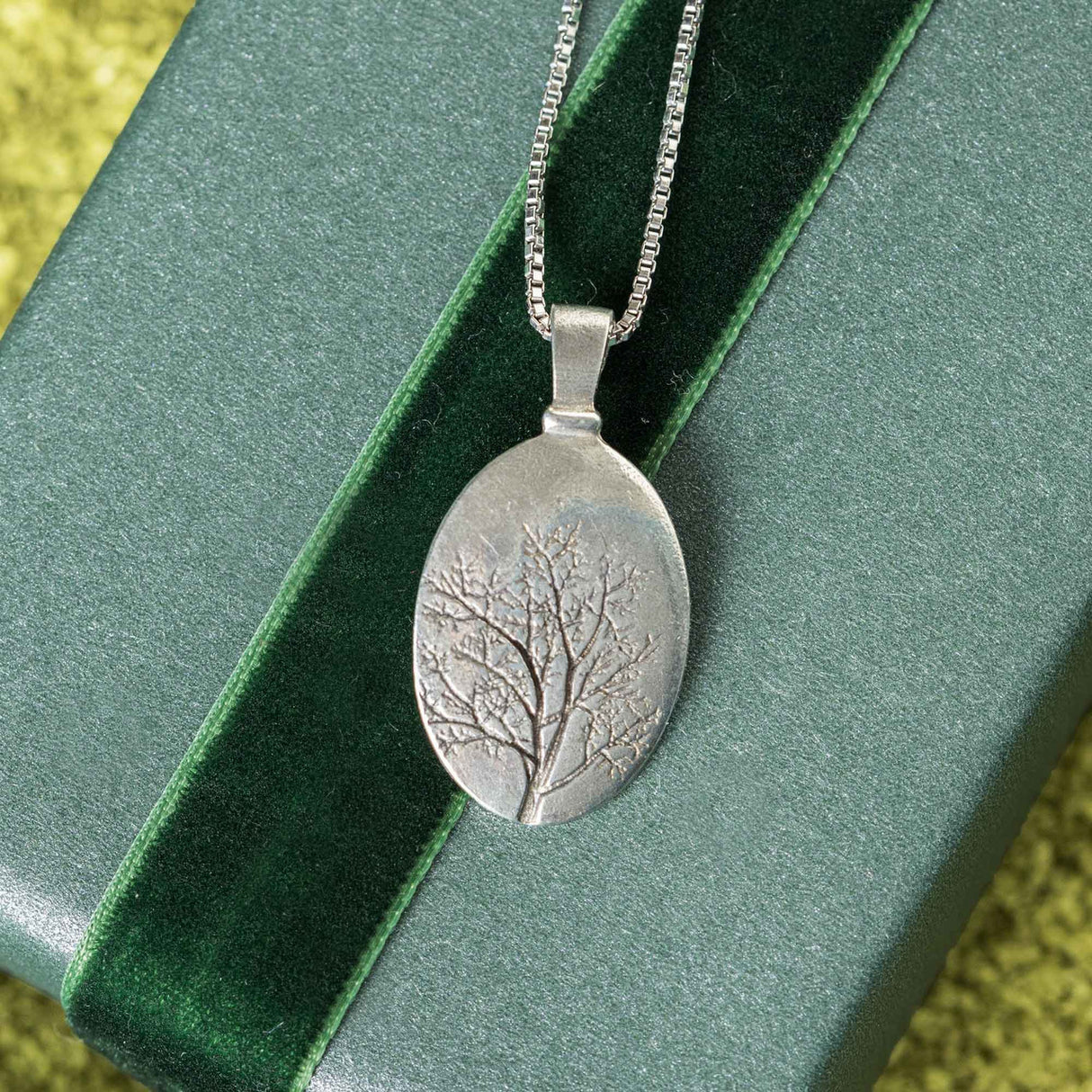 a pewter oval pendant with finely crafted engraving of a tree with delicately detailed branches laid over a light green box