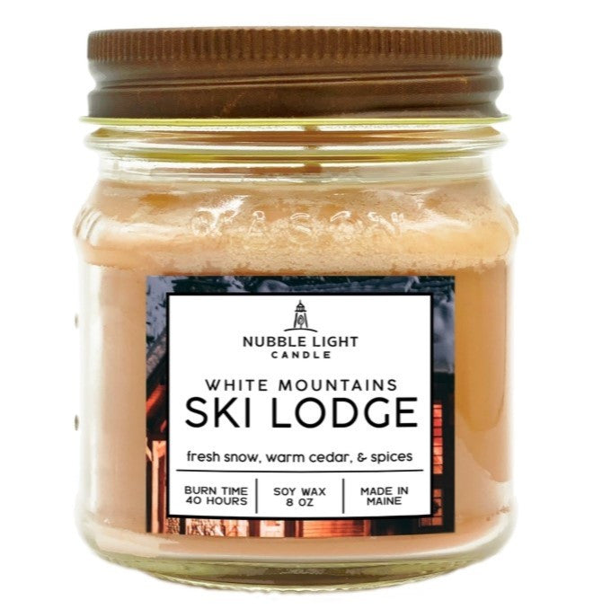 a candle in a clear glass mason jar with tan wax and a label that reads "White Mountains Ski Lodge" against a white background
