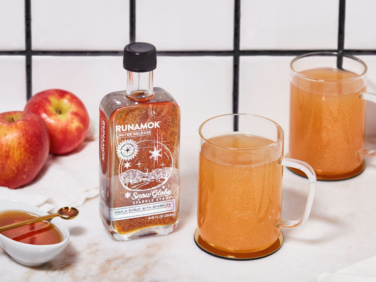 snow globe sparkle syrup on a countertop next to apples and clear mugs showing a sparkly cider drink