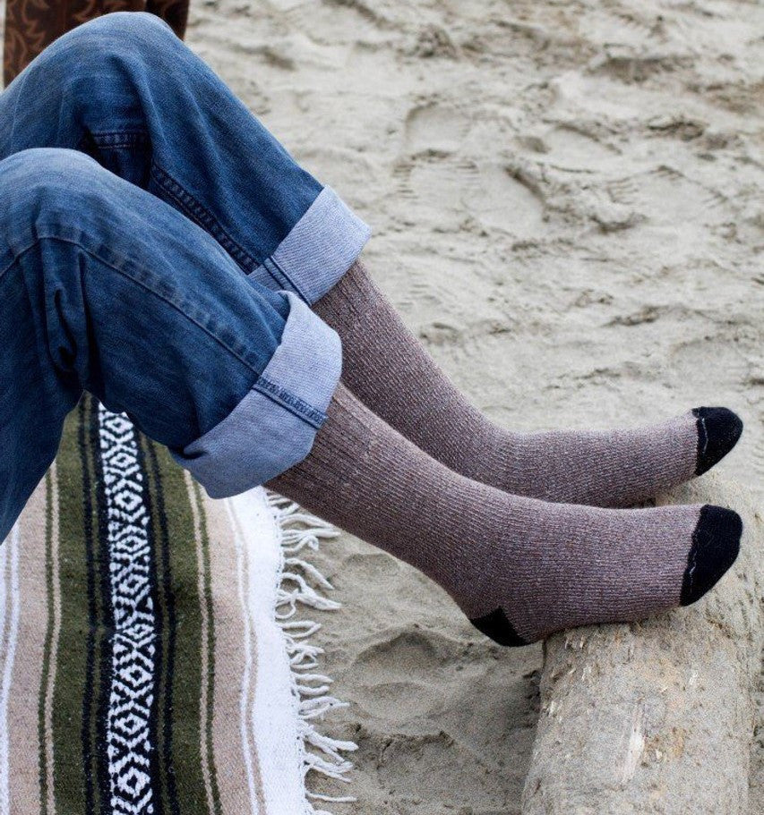a pair of legs wearing rolled up jeans and cocoa brown alpaca socks resting their feet on a log with beach sand visible in the background