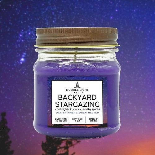 a candle in a clear glass mason jar with purple wax and a label that reads "Backyard Stargazing" against a starry night sky