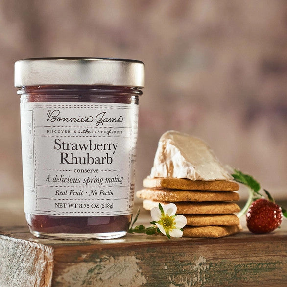 Bonnie's Strawberry Rhubarb jam jar with an elegant white label and silver jar top on a table next to a stack of crackers topped with a soft cheese and herbs