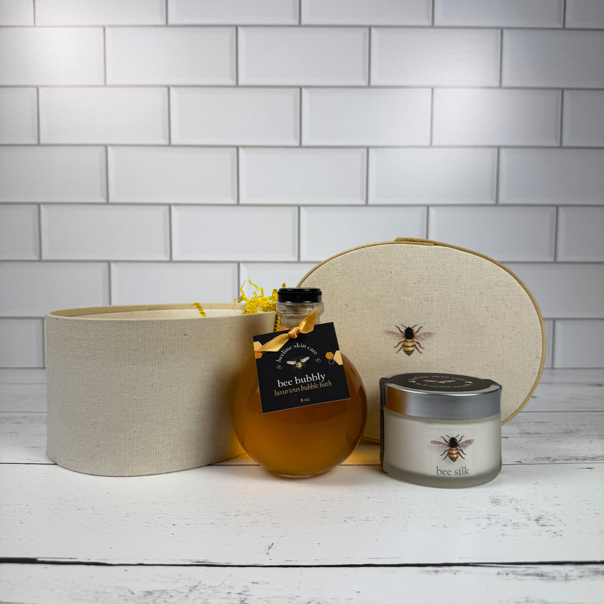 a spherical glass bottle of honey colored bubble bath with a jar of Bee Silk creme in front of an elegant off-white hat box with a bee printed on the top