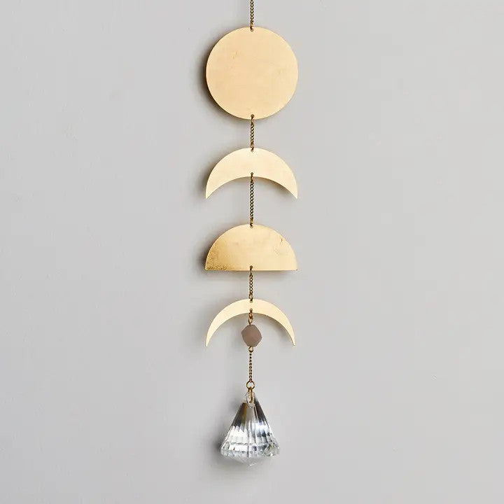 an elegant and minimal suncatcher in brass featuring a full circle representing the full moon with cresent and half moon shapes below and a moon stone crystal pendant hanging at the bottom