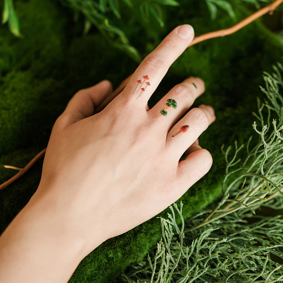 close up of a model's hand with colorful mushroom tattoos applied between their knuckles to show the smaller size of some of the tattoos included in the set