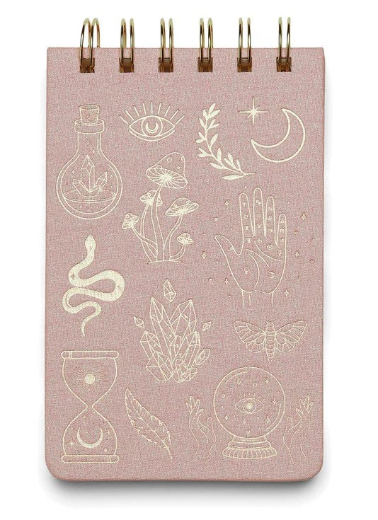 a pale pink notebook with spiral wire top binding and gold foil illustrations of mystical themed icons including an hourglass, crystals, mushrooms, palmistry hand, crescent moon, feather, snake, and more
