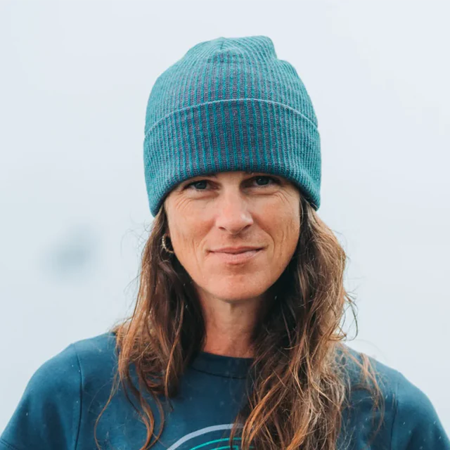 A woman with long brown hair wearing a teal ribbed knit beanie and a matching teal sweatshirt, photographed outdoors against a soft, overcast light gray background.
