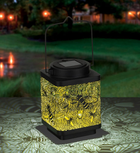 On a table in a garden at twilight an illuminated, cube shape lantern with intricate design of bees in flight carved out of the sides of the lantern so the light casts shadows of the bee design on the surface surrounding the lantern.