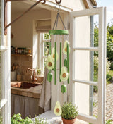 Green avocado-themed wind spinner hanging in front of an open white window, with avocado slices, green bars, and seed details, set in a bright kitchen and garden environment.