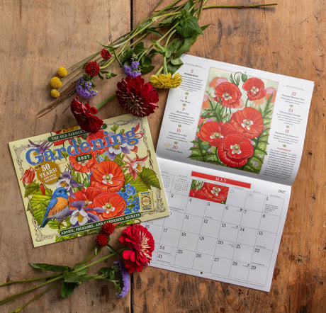 A gardening calendar and its illustrated cover box are arranged on a rustic wooden surface with colorful flowers. 
