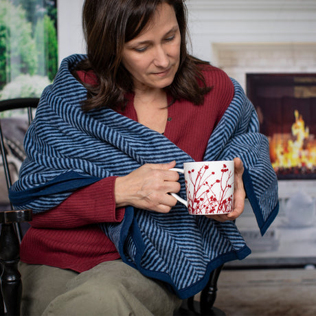 a seated woman looks down at her winterberries mug while wrapped in the Harborview Herringbone Blanket