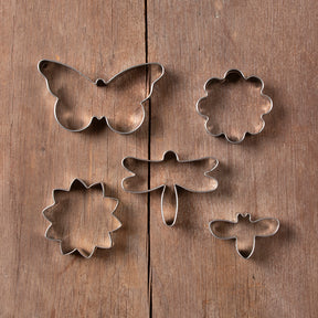 Set of metal cookie cutters shaped like a butterfly, flower, sun, dragonfly, and bee on a wooden surface.