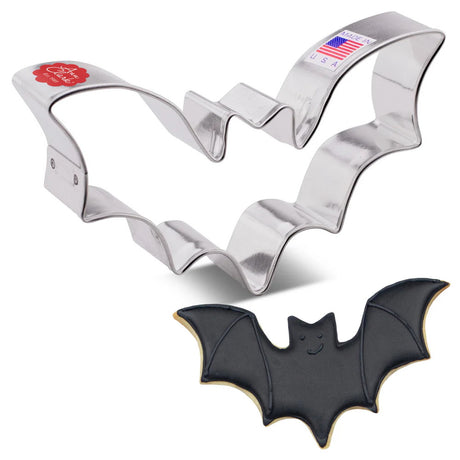 A tin cookie cutter in the shape of a flying bat with wings spread next to a fully decorated bat cookie with black icing, wing detail, and a cute smiling face
