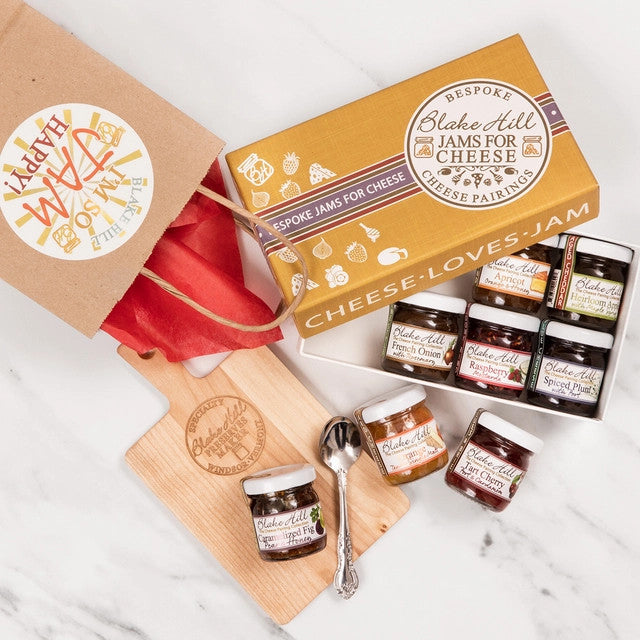 top down view of the cheese loves jam box on a marble countertop with the box partially open revealing jams in the box along with 3 jars outside of the box on a cheese board with a spoon