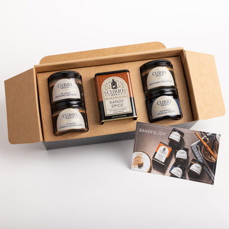 A curated gift box of Curio Spices containing four spice jars and a box of Kandy Spice, displayed in an open brown cardboard box with a 'Baker's Joy' card on a white background.